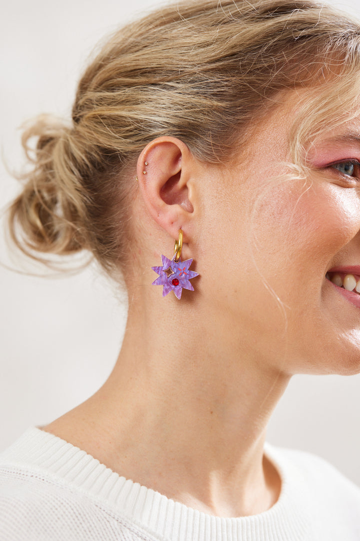 Purple star  earrings with twinkling motifs and a dazzling red crystal inlay hanging on a gold hoop pictured on a model 