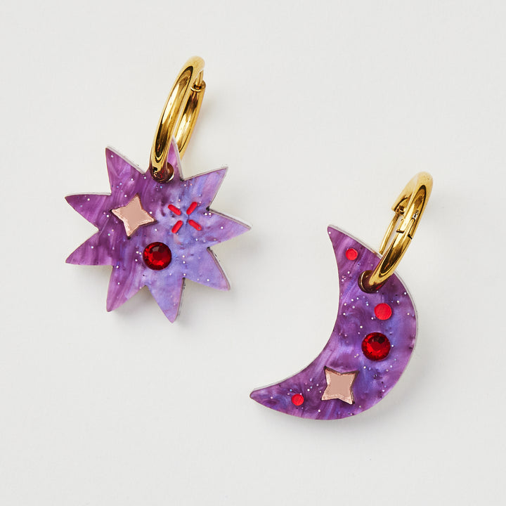 Purple mismatched star and crescent earrings with twinkling motifs and a dazzling crystal inlay hanging on a gold hoop 